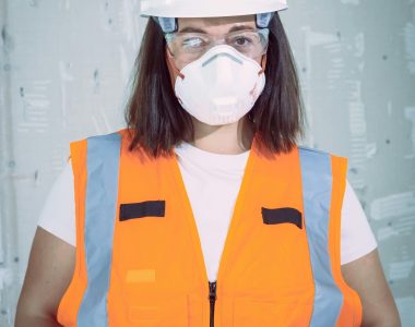 Female engineer in safety gear with hard hat and N95 mask indoors.