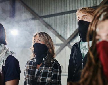 A diverse group standing indoors wearing face masks, conveying a sense of mystery or protest.